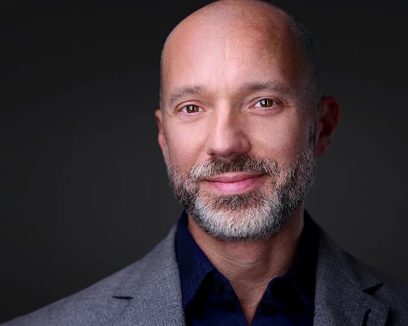 Professional headshot of a bald man with a short gray beard against a dark background